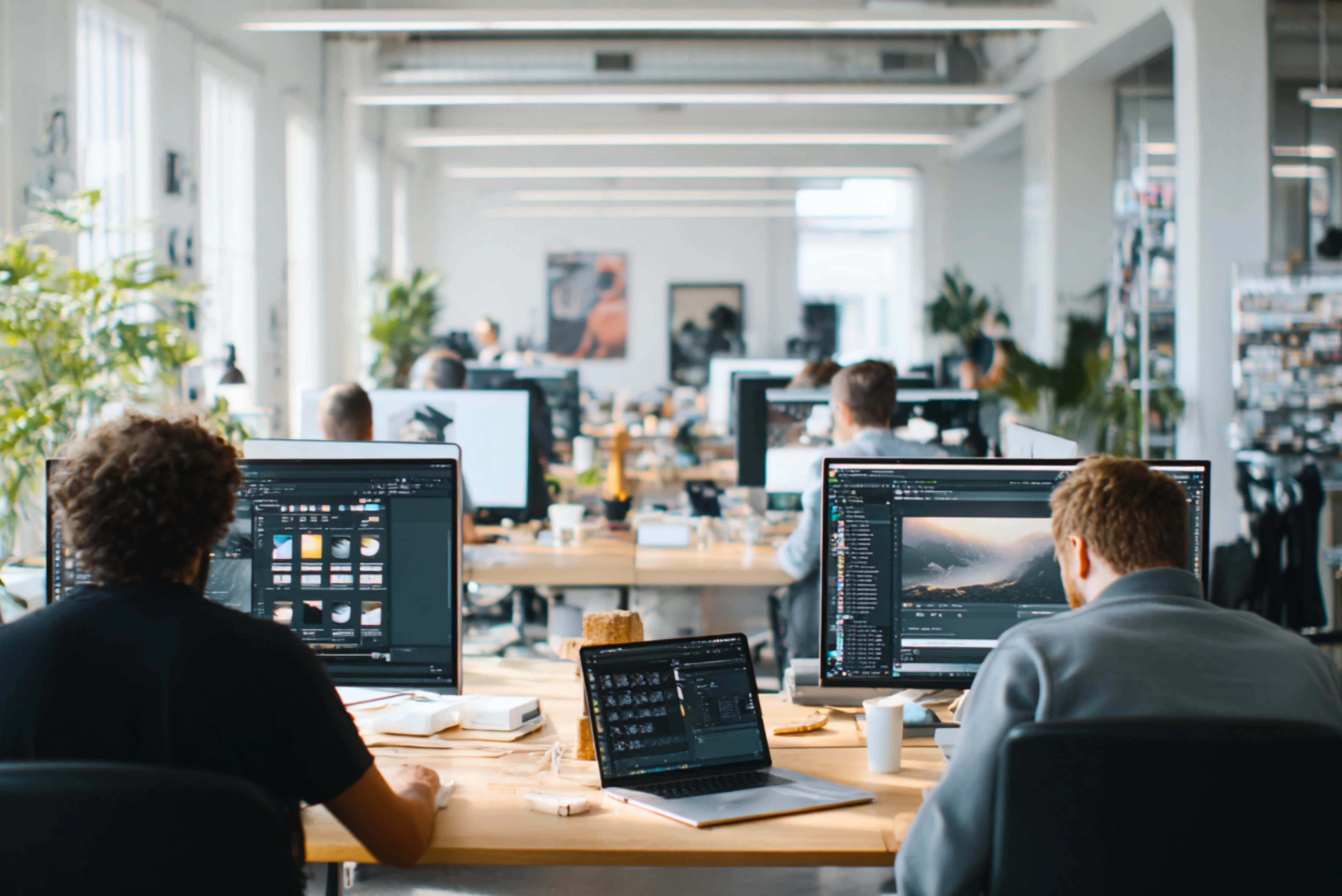 People working in a modern open-plan office, focusing on graphic design and video editing on large computer monitors and laptops.