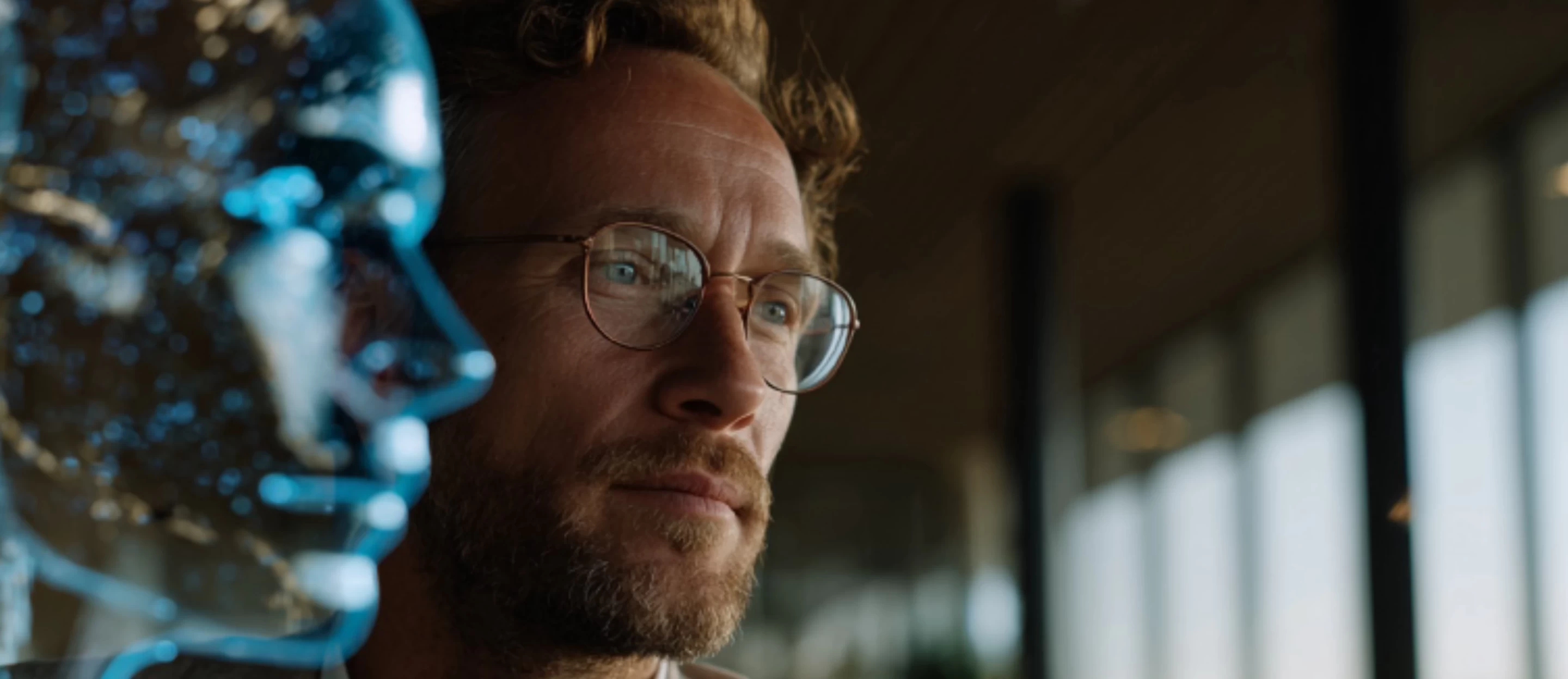 A man wearing glasses looks thoughtfully at a translucent blue AI face model beside him.